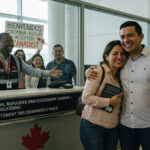A happy migrant couple at a Canadian airport celebrating their asylum acceptance with family waiting behind glass.